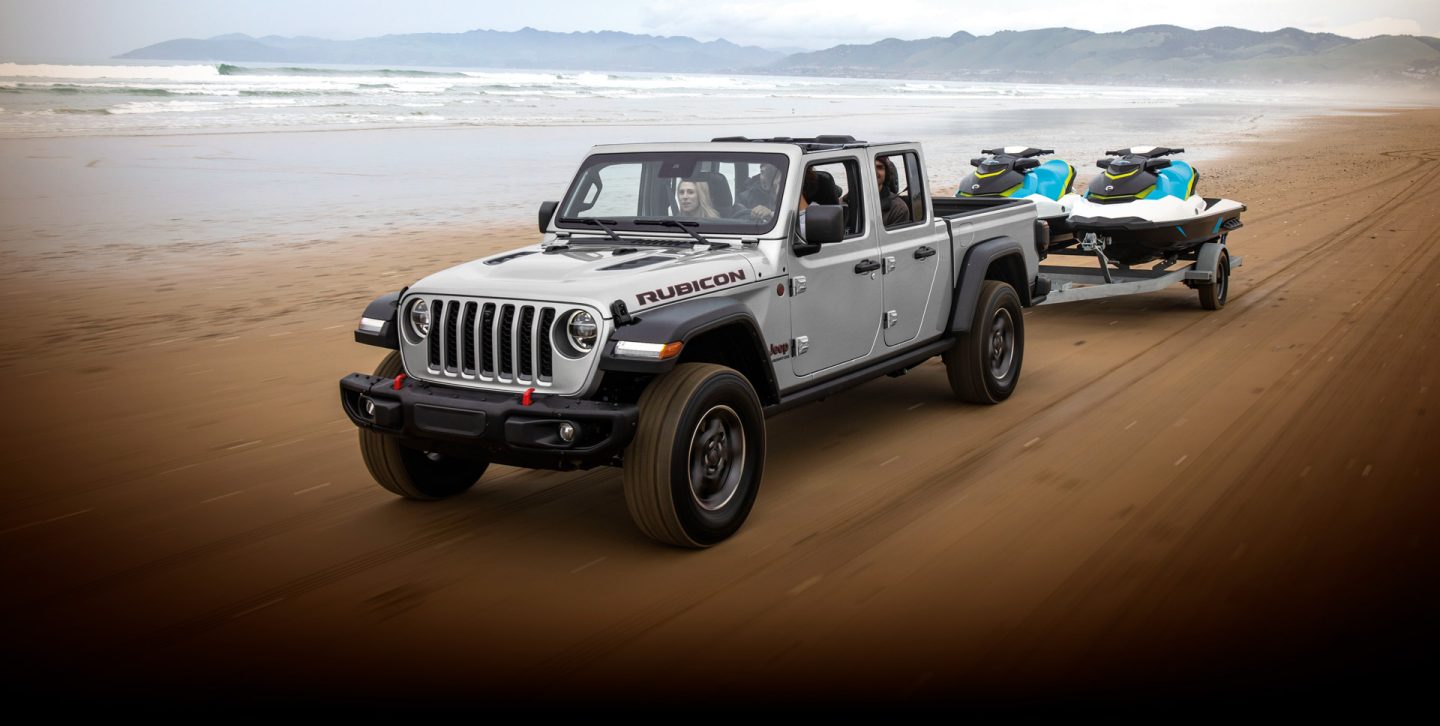 A 2022 Jeep Gladiator Rubicon towing two jet skis as it is driven along a beach near the water.